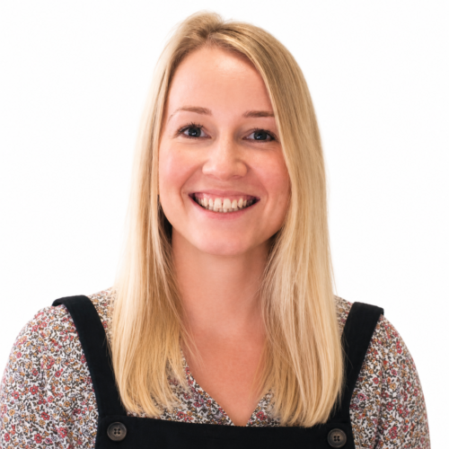 Smiling woman with long blonde hair, facing the camera in a clean, well-lit headshot against a plain light background, wearing a floral top under a black pinafore-style dress.