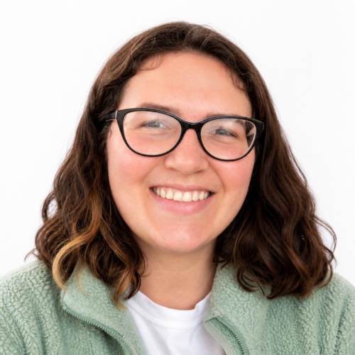 Smiling woman with shoulder-length wavy dark hair and glasses, wearing a light green fleece, in a clean, well-lit headshot against a plain white background.