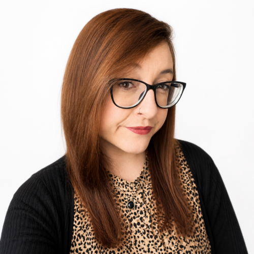 Smiling woman with long auburn hair and glasses, wearing a patterned top and black cardigan, in a clean, well-lit headshot against a plain white background.